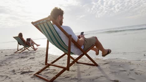 vista lateral de un joven caucásico sentado en una tumbona y usando un teléfono móvil en la playa 4k