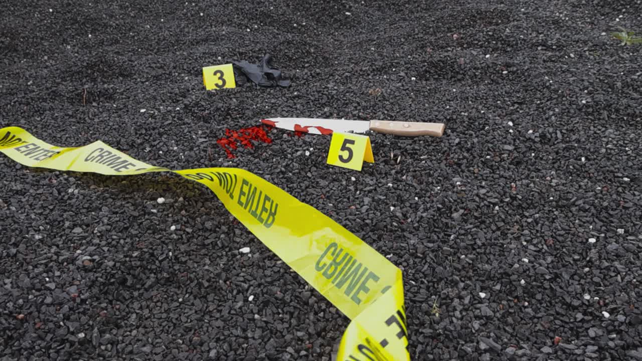 Yellow crime scene police line crime scene tape and crime markers next to evidence like bloody kitchen knife and a black nylon glove on dark gravel ground outdoors duriing day time. Wind movement