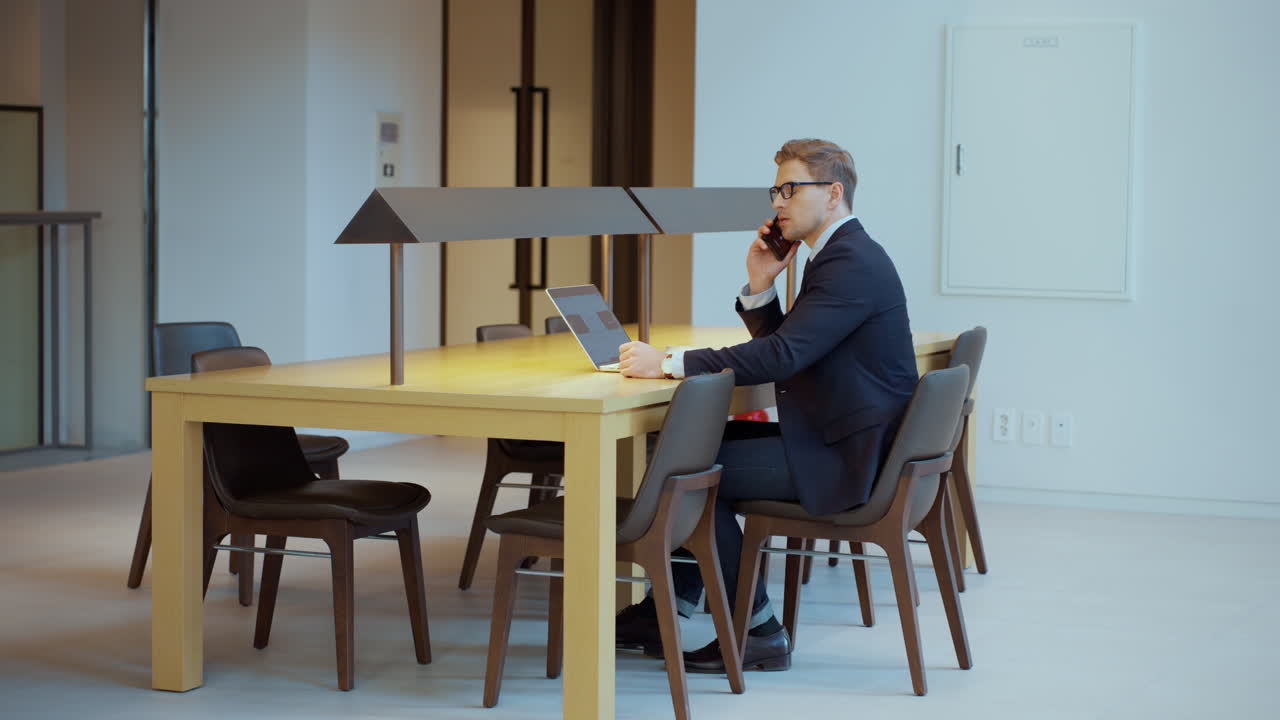 Young businessman having phone conversation sitting alone in spacy offce meeting room - revealing full shot