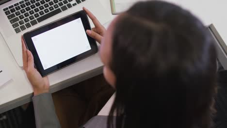 Mujer-De-Negocios-Birracial-Sentada-En-El-Escritorio,-Usando-Una-Tableta-Con-Espacio-Para-Copiar-En-Una-Oficina-Moderna