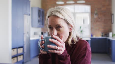 Thoughtful-senior-caucasian-woman-in-living-room-sitting-on-sofa,-drinking-coffee