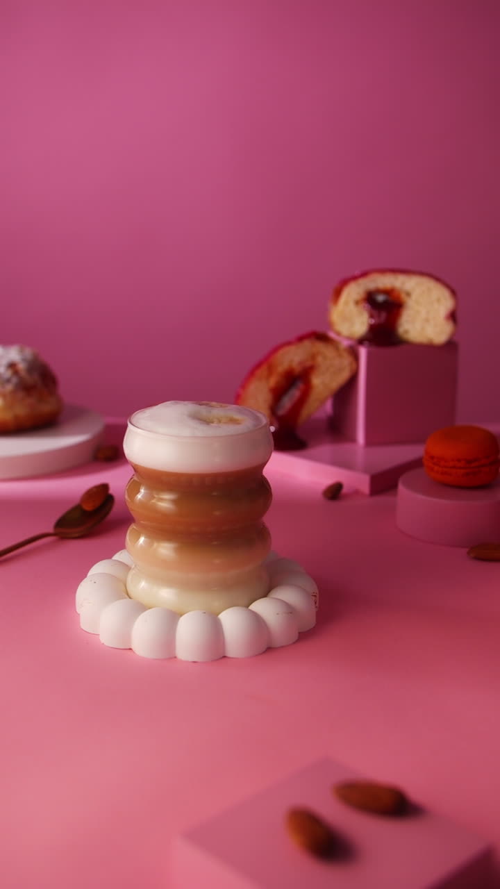 Slowmotion shot of several almonds falling in front of a layered latte glass. Pink background, styled with a Berliner donut, sliced cake, orange macaron, and golden spoon on a minimal dessert setup