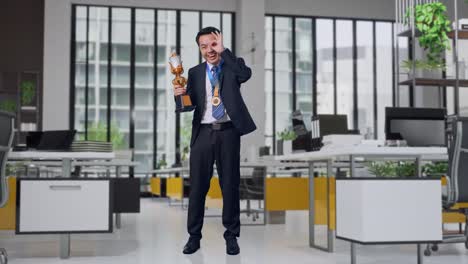 full body of asian business man in a suit and tie with a gold medal and trophy showing okay hand sign over eye and smiling to camera as the first winner in office