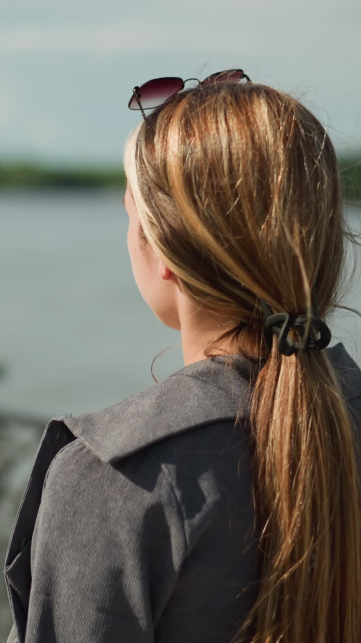 다리 울타리 근처에서 산책하는 젊은 여자의 뒷면, 머리에 선글라스를 고, 바람이 그녀의 머리카락을 통해 불고, 배경에는 강의 흐릿한 시야가 있습니다.