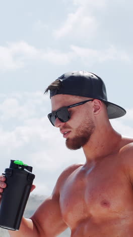 bodybuilder man drinks water from a bottle on a background of the sky with clouds. shirtless athlete in sunglasses and a black cap. vertical video