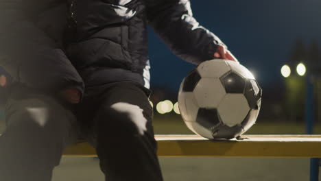 close up of someone seated on a bench with a worn soccer ball beside them, the wind moves the ball slightly, and the person steadies it to prevent it from falling