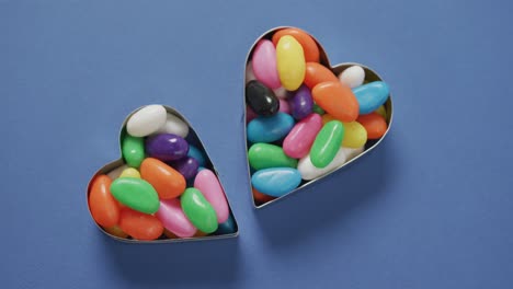 video of overhead view of multi coloured sweets in two heart shapes over blue background