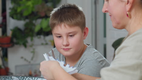 close view of boy staring attentively at paper craft while mother carefully carves paper with scissors, both focused on creative project at home, indoor scene with blurred plants in background