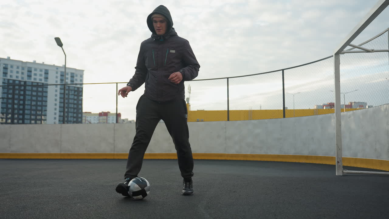 homem de capuz praticando exercícios de futebol em uma arena esportiva, demonstrando trabalho de pés e controle de bola sob a luz do dia, o fundo mostra poste de lâmpada, edifícios residenciais urbanos e poste de gol em uma quadra cercada