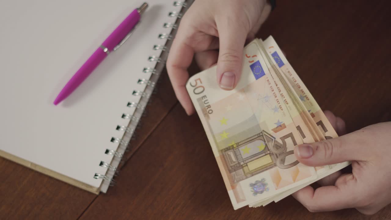 Mani che contano banconote accanto a un quaderno a spirale e una penna su una scrivania di legno, ideale per concetti di budgeting, finanza, risparmio e contabilità.
