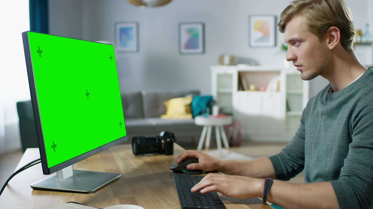 un bel giovane fotografo lavora su un computer personale con uno schermo verde mentre è seduto alla sua scrivania nell'accogliente appartamento.