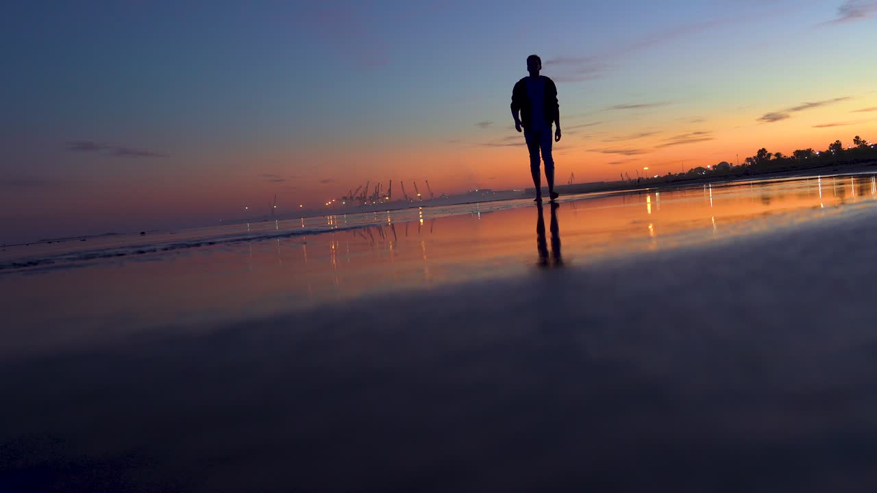 Man walks on the sand barefoot at sunset on a beautiful beach, his footprints on the wet sand and the stagnant water of the sea waves in backlight