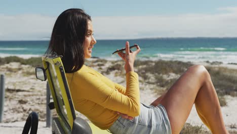 mujer caucásica acostada en un buggy de playa junto al mar hablando en un teléfono inteligente