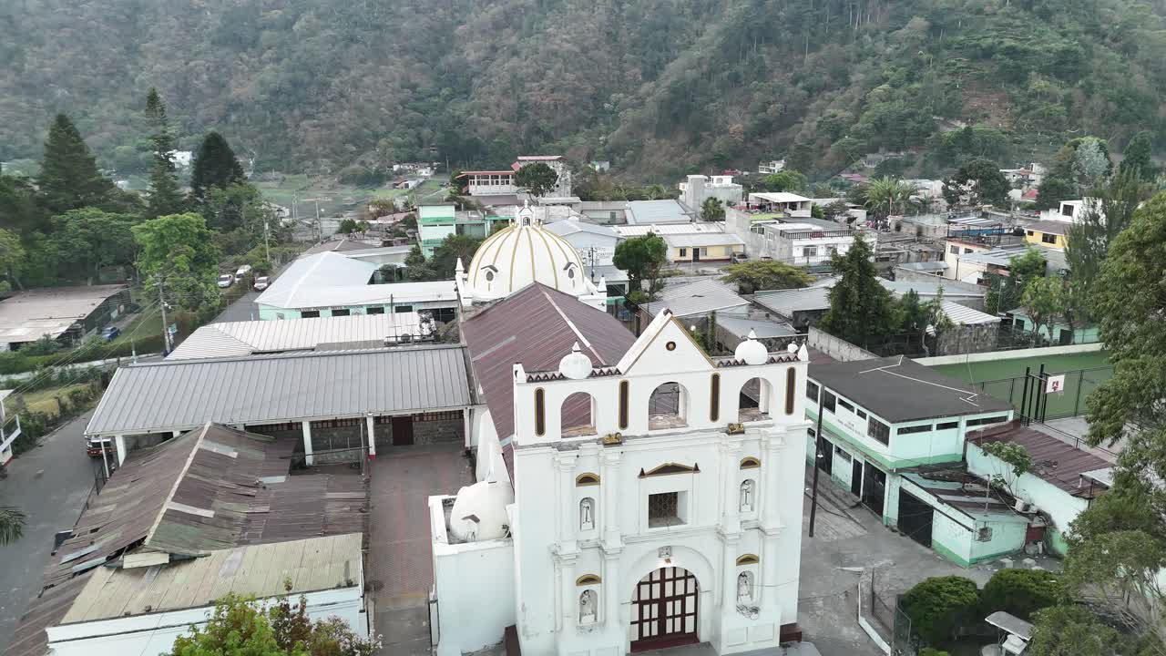 Drone aerial view over San Lucas Guatemala captures a historic church, tropical neighborhoods, palm trees, and forested volcano mountains rising into a bright sky for a stunning landscape
