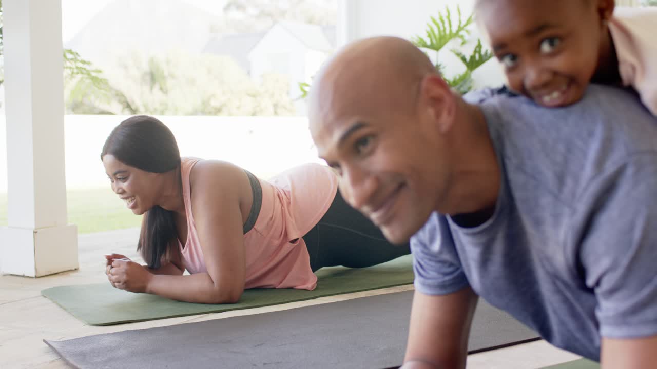 African American man planking on patio reacting to partner's smile as child climbs his back bonding