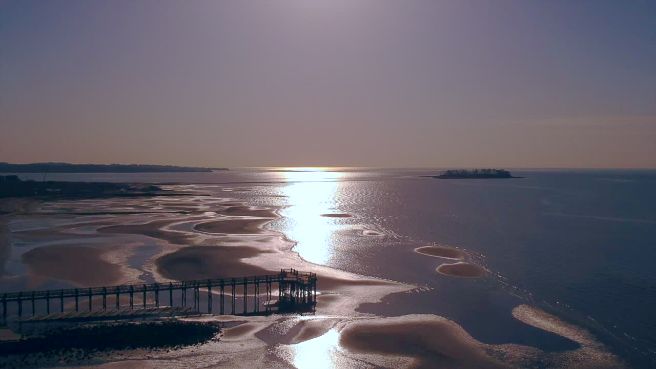 Drone descending over a beach at sunrise with a pier and an island in the background
