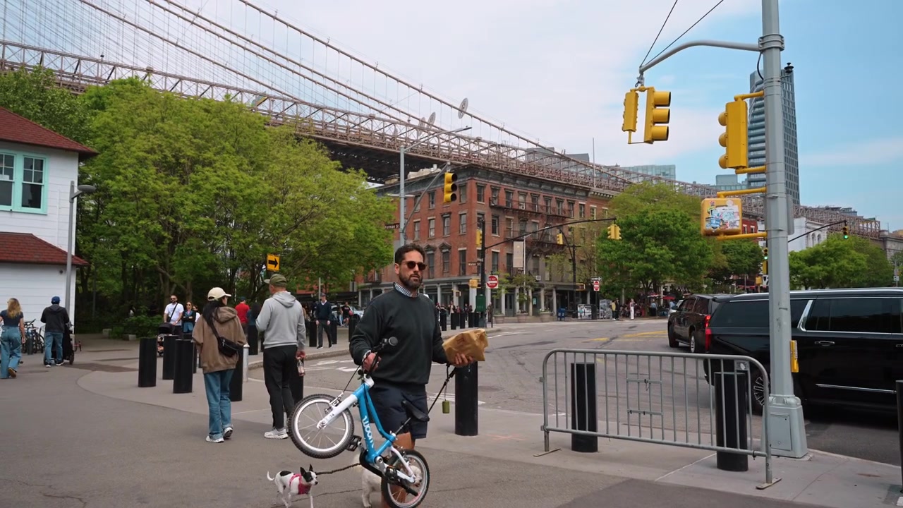 New York, USA, 19 maj 2026: Gatuvy dagtid över DUMBO-korsningen, Brooklyn, New York. Människor korsar vägen nära trafikljus med tegelbyggnader och grönskande lövverk under brospannet