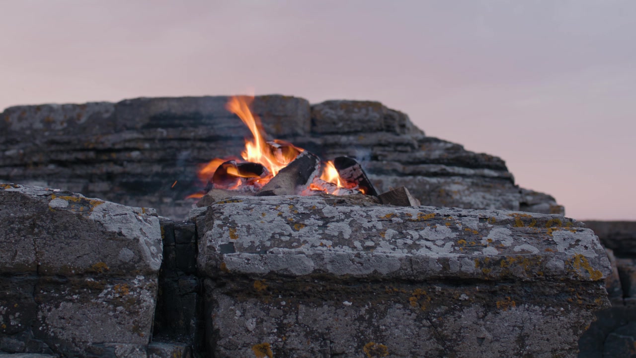 Caldo falò che brucia tra aspre rocce costiere al tramonto, con fiamme incandescenti che creano una suggestiva scena all'aperto in riva al mare