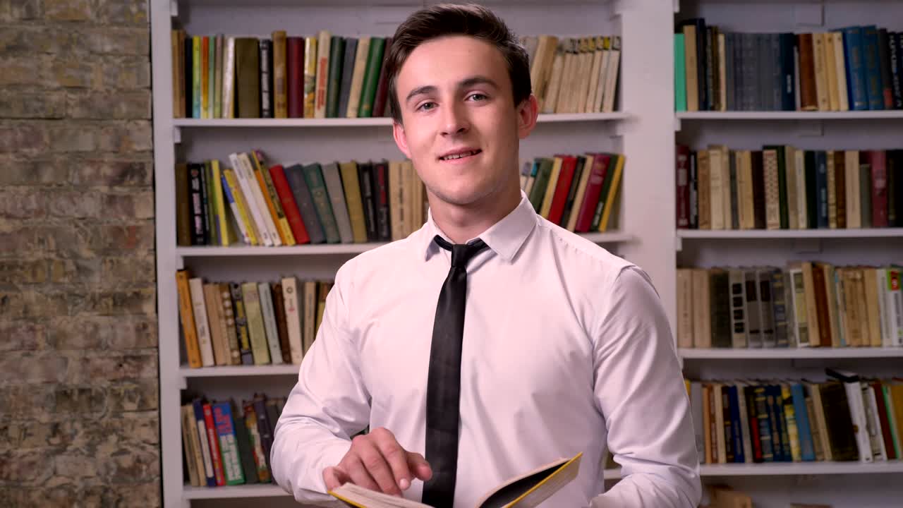 joven empresario está buscando información en el libro en la biblioteca, mirando a la cámara, concepto de relajación