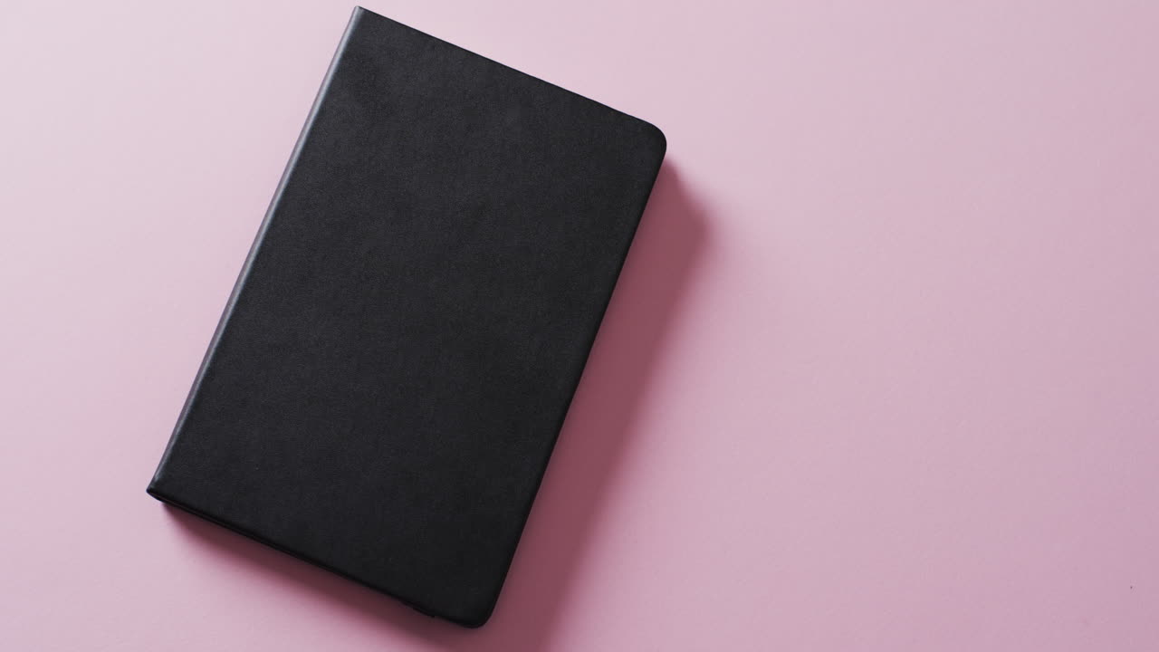 Close up of closed black book with copy space on pink background in slow motion