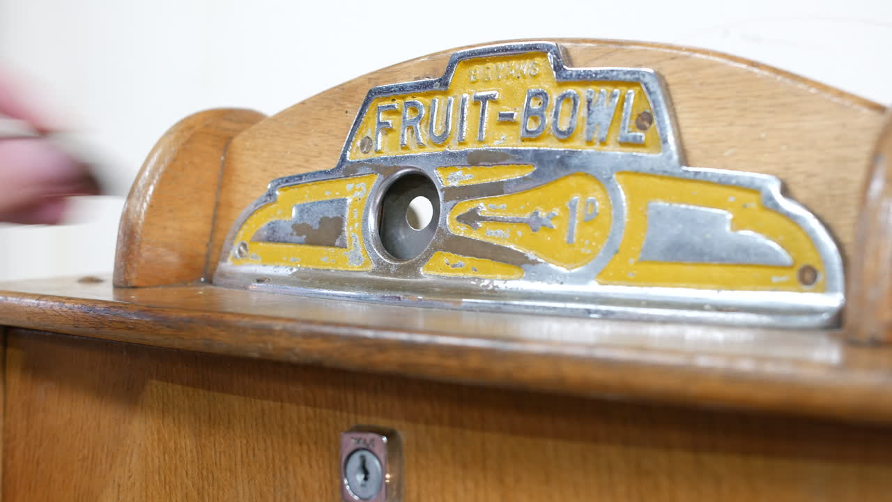 A hand inserting a coin into a vintage Fruit-Bowl machine is interacting with a metal coin slot mounted on a wooden panel labeled with retro embossed graphics