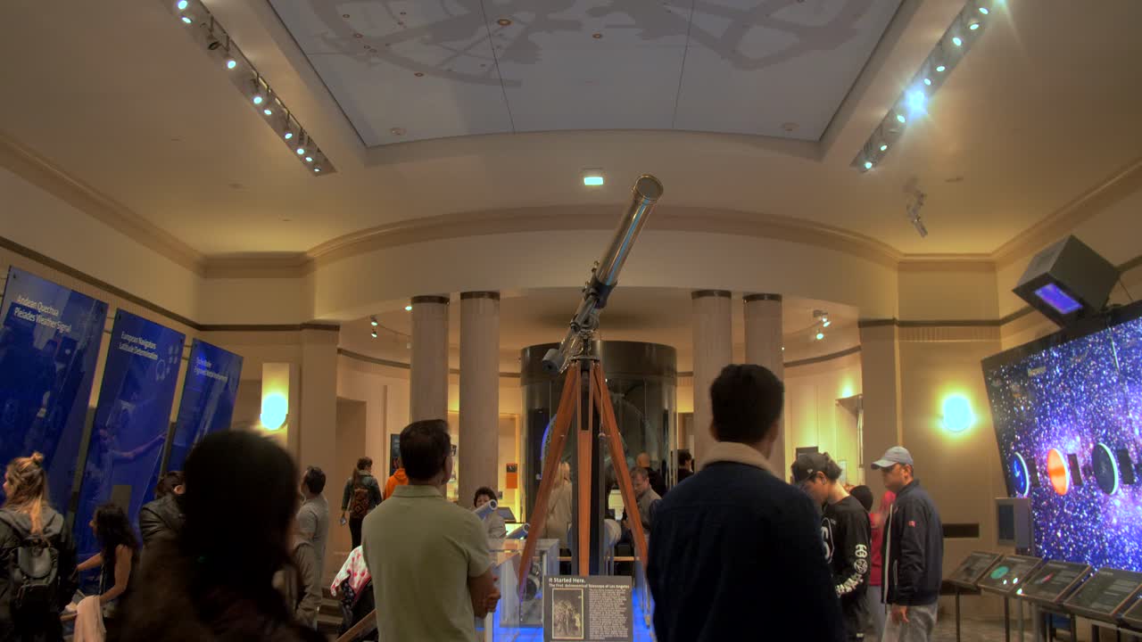 Griffith Observatory in Los Angeles, showing an old Telescope from ancient times with gold exterior being viewed by lots of museum visitors.