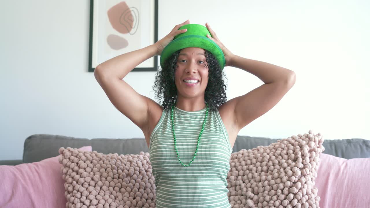 Donna afroamericana su un divano che sistema gli occhiali da sole, scambiando un cappello verde e occhiali a forma di trifoglio con una macchina fotografica.