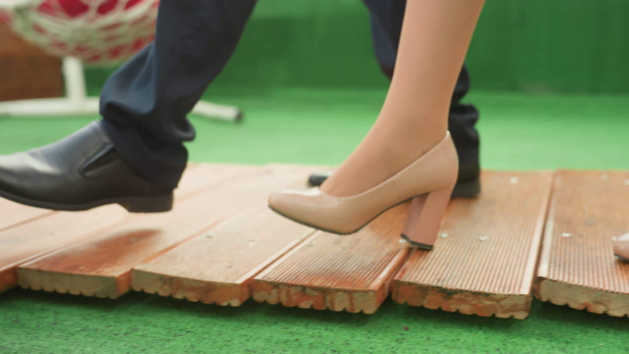 casal caminhando em deck de madeira com saltos, close-up intimista dos pés sob a mesa vermelha com jantar posto, mulher usando saltos bloco e homem com sapatos sociais de couro preto pisando em tábuas desgastadas