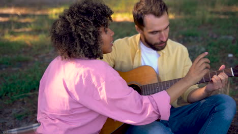 una mujer le enseña a un hombre a tocar la guitarra. una pareja de excursionistas felices acampando en el bosque. una joven negra y un hombre caucásico.
