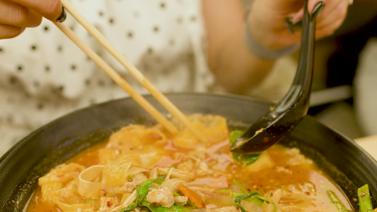 Hand lifts tofu with chopsticks and spoon from vibrant spicy noodle soup under warm lighting