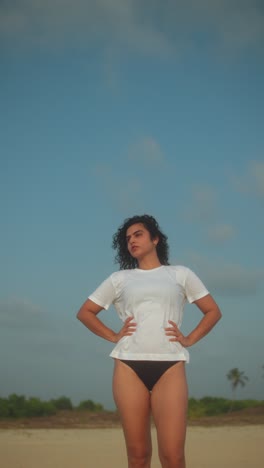 indian woman in white t-shirt and bikini bottoms stands confidently on sandy beach