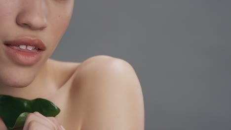 Primer-Plano-Retrato-Hermosa-Mujer-De-Raza-Mixta-Sonriendo-Acariciando-La-Cara-Con-Hojas-Tocando-Una-Tez-De-Piel-Suave-Y-Saludable-Disfrutando-De-La-Belleza-Natural-Del-Cuidado-De-La-Piel-Sobre-Fondo-Gris