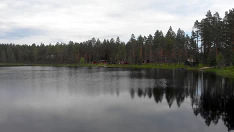 Drone-flight-through-pine-tree-forest-towards-a-lake-with-some-wooden-cabins-located-at-the-edge-in-Dalarna-county,-Vansbro-municipality-in-Sweden