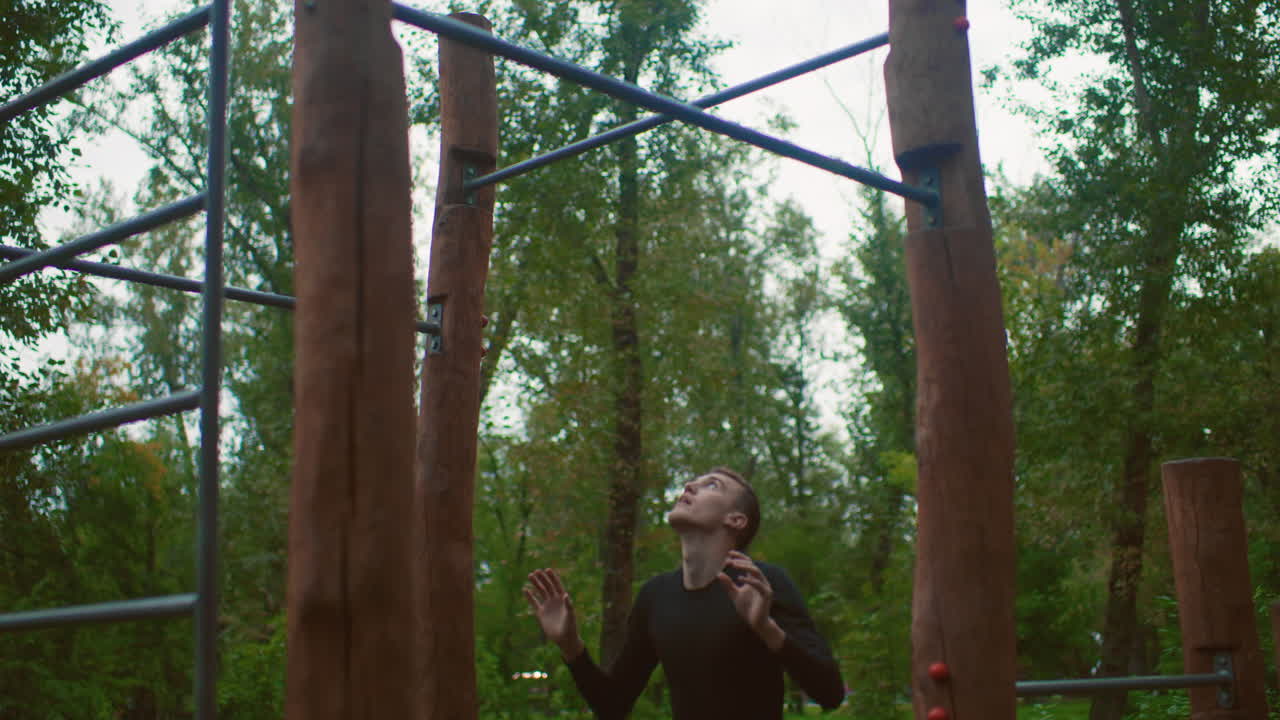 man struggling on outdoor pullup bars, wooded training area with wooden posts and metal crossbeam, performing hanging negatives and controlled descents, focused strain, overcast sky, solo practice