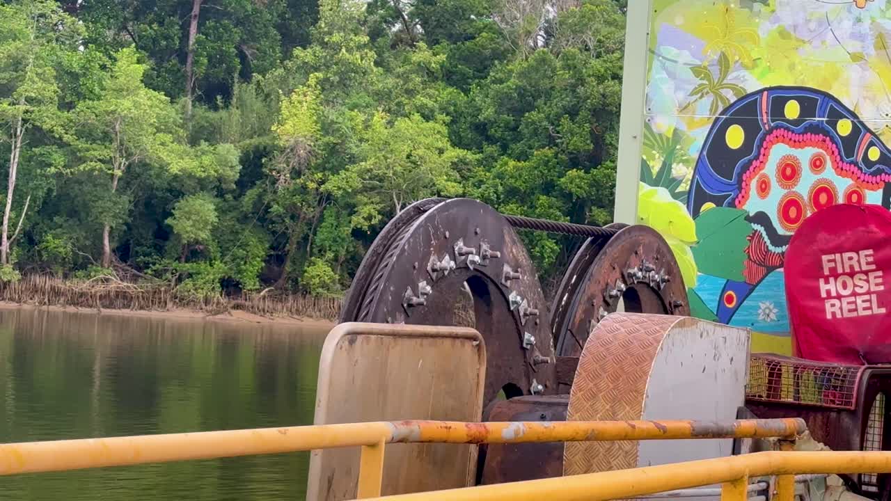 Rusty ferry cable wheels operate beside river, lush rainforest background, overcast daylight, steady shot