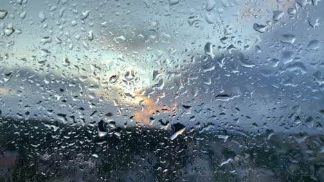 Primer-Plano-De-Las-Gotas-De-Lluvia-En-La-Ventana,-Puesta-De-Sol-Borrosa-En-El-Fondo