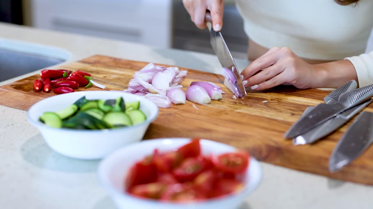 A person slices shallots on a wooden board surrounded by fresh vegetables in a bright kitchen setting
