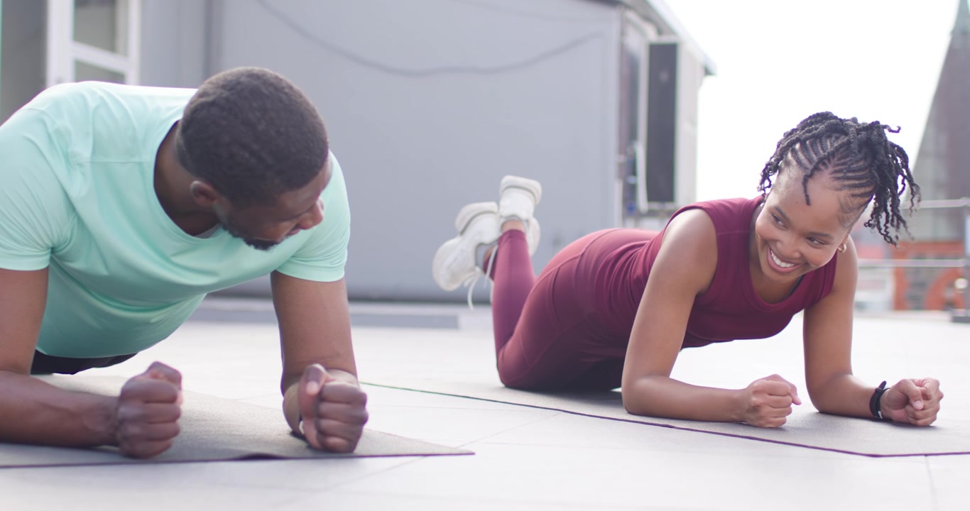 African friends planking on rooftop mats in workout wear, woman entering, smiling and encouraging