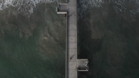 AERIAL-birds-view-flight-over-pier-with-waves-crashing-into-beautiful-shore-line