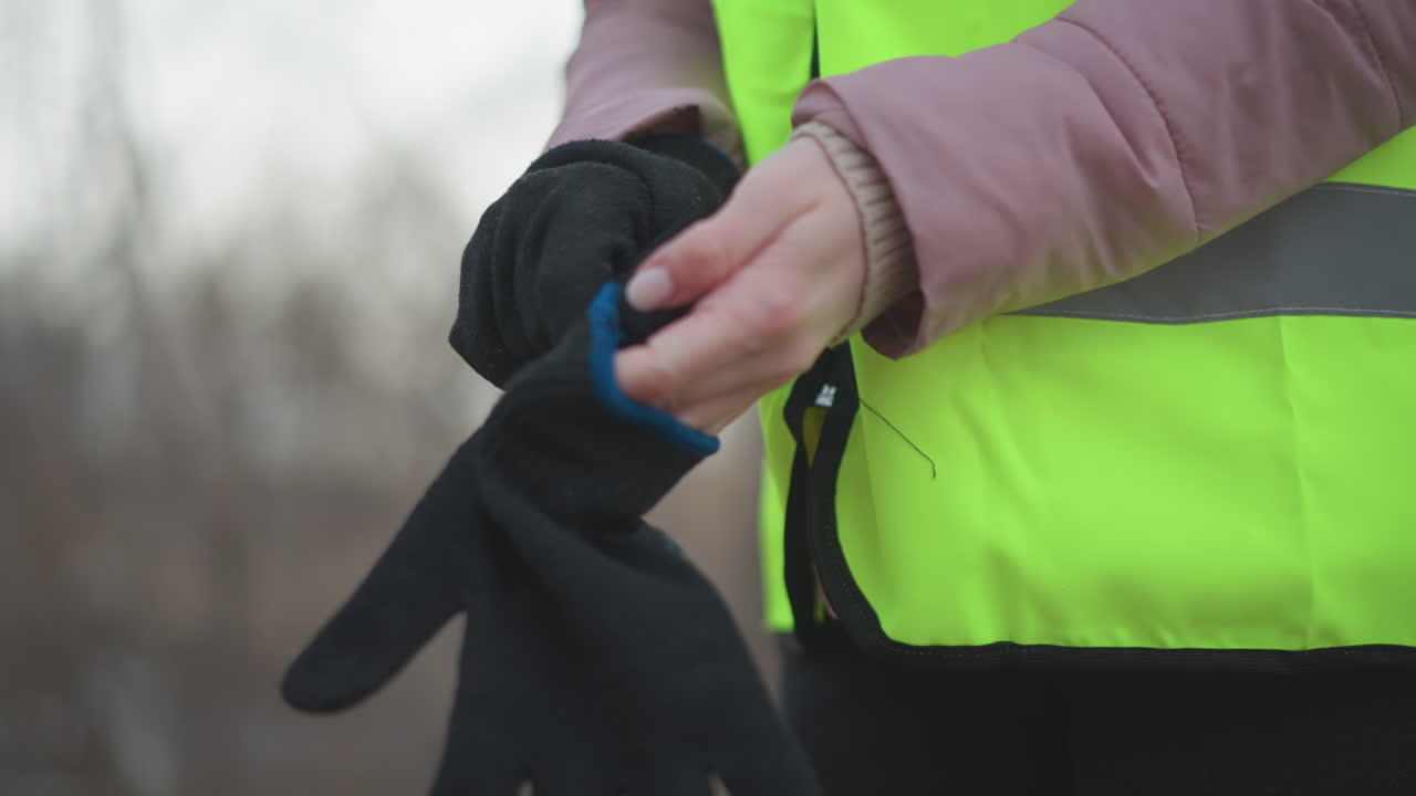 Close-up of person in neon yellow reflective safety vest and pink padded jacket putting on black knitted gloves outdoors during cold weather, preparing for outdoor work or safety task in winter