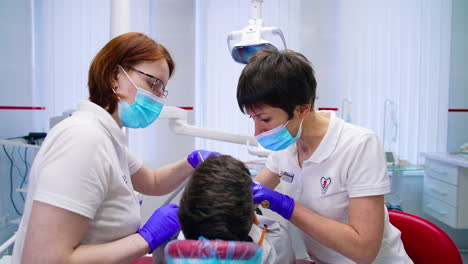 Patient in dental chair treating teeth. Female specialist and a nurse doing dental procedure to boy's teeth in modern stomatology office. Rear view.