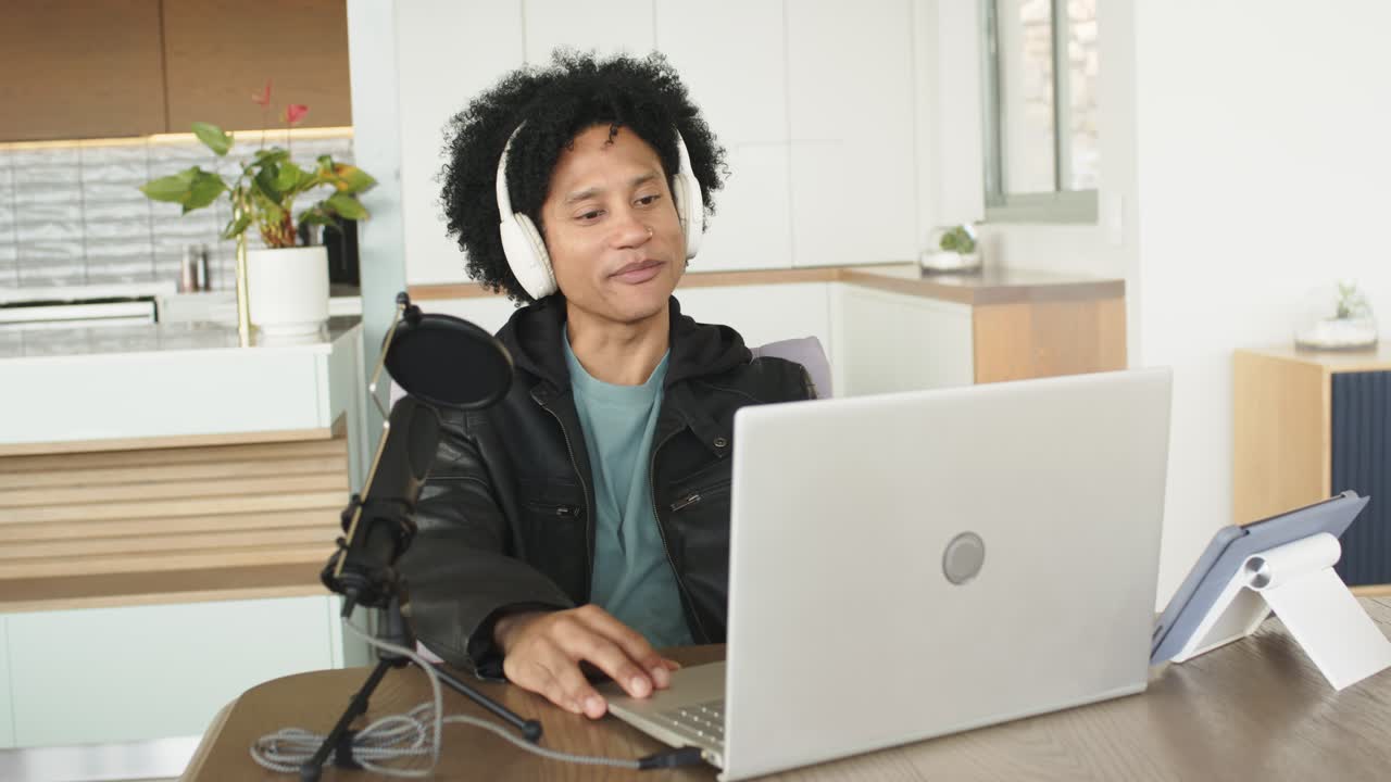 African American man typing on laptop, recording with microphone at kitchen table with headphones