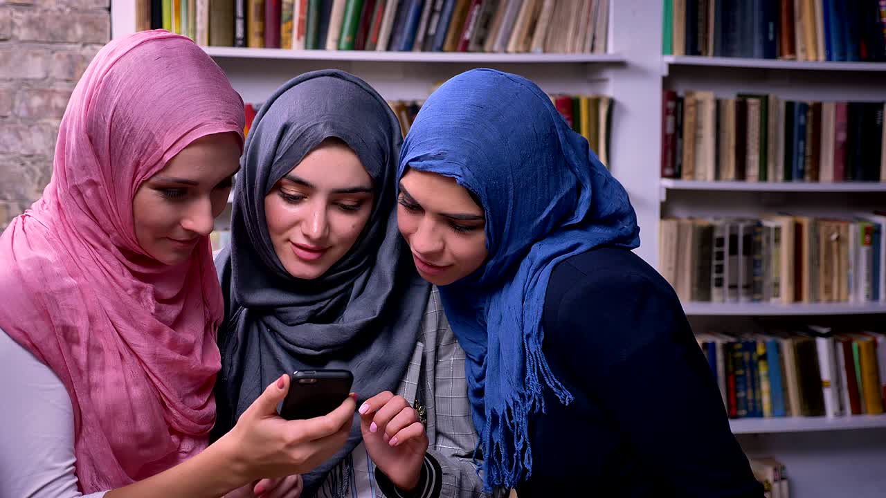 потрясающие девушки в хиджабе смотрят на телефон и улыбаются, стоя вместе в библиотеке, ближневосточная иллюстрация