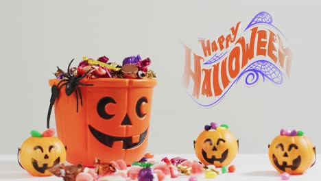 animation of halloween text over carved pumpkin bucket with sweets on grey background