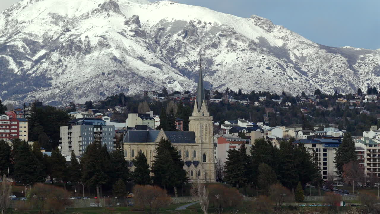 Left-tracking drone clip frames historic Bariloche cathedral against the massive, snow-covered Cerro Catedral mountain