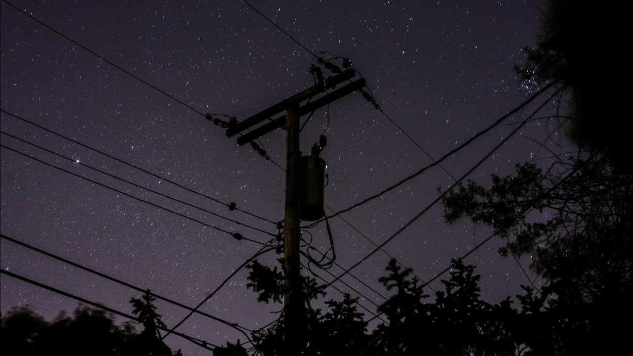 A time-lapse of the stars rotating in the sky behind a silhouetted suburban telephone pole and power lines and trees in the night sky