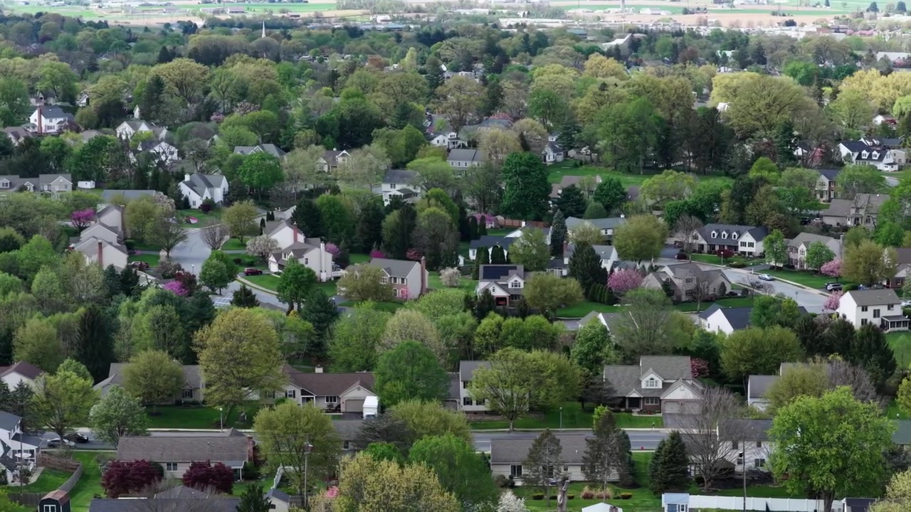 Aerial view above green suburban neighborhood in USA with modern homes, tree lined streets and peaceful residential housing surrounded by fresh spring foliage and gardens. Wide shot.