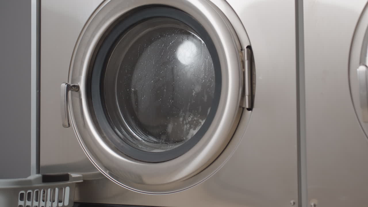 Round view stainless front load laundry machines rolling clothes with water splashes behind glass door, nearby floor basket, busy wash cycle in progress, laundromat scene with metal drum and seal