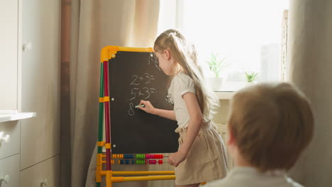 Girl-writes-math-problems-for-brother-playing-school-at-home