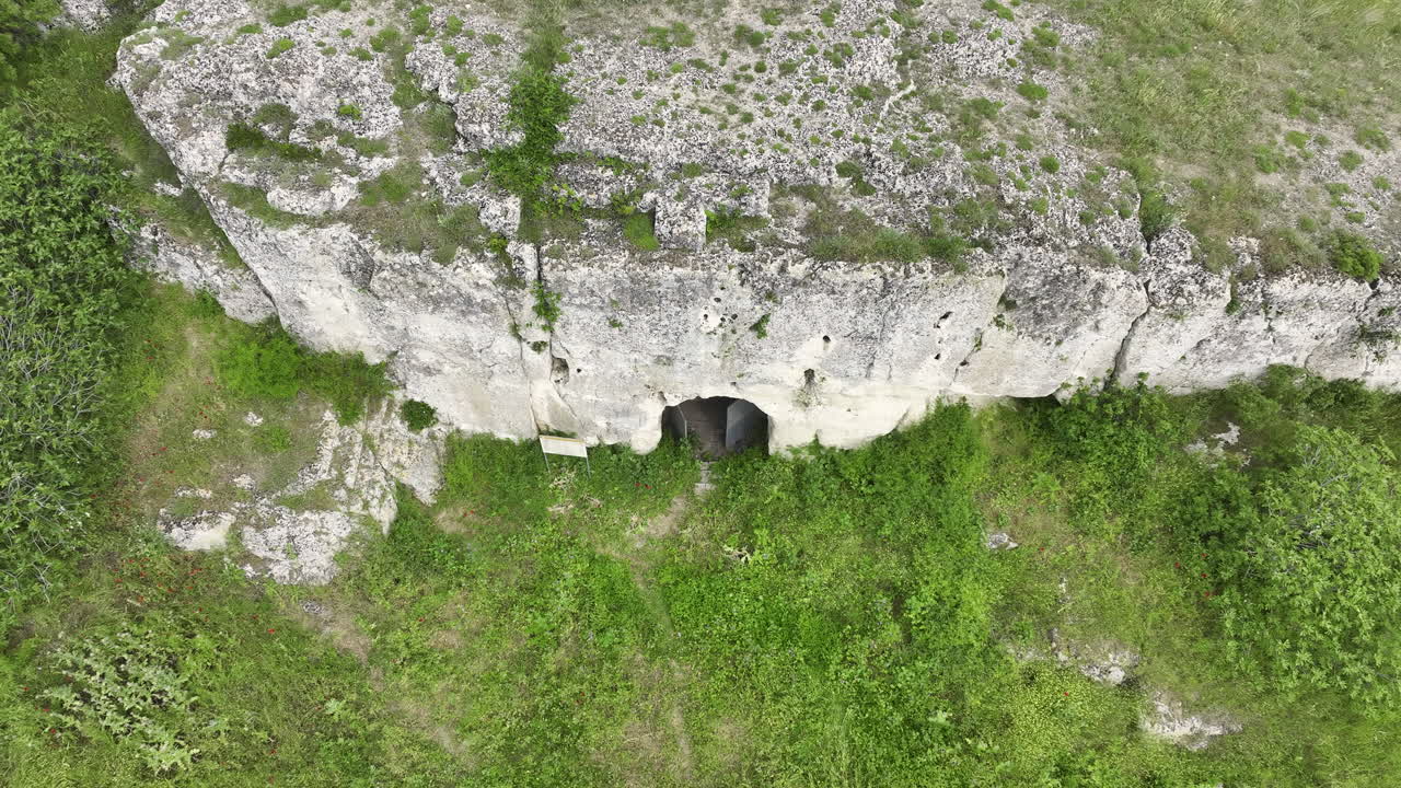 Pull-back drone shot revealing the arched doorway of a rock-hewn medieval cave church carved into a limestone cliff, surrounded by lush spring greenery and meadows under soft, overcast light